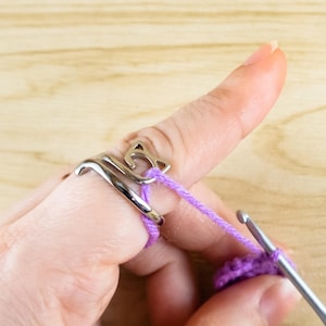 Puede incluir: Un anillo guía de hilo plateado con forma de gato, usado en un dedo, con hilo morado que lo atraviesa. Un ganchillo se ve en la esquina inferior derecha, trabajando en un proyecto de ganchillo morado. El fondo es una superficie de madera clara.