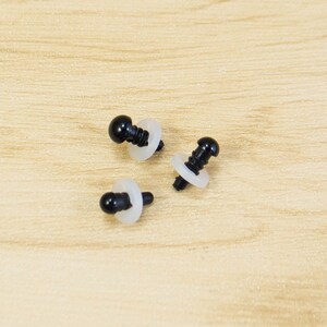 May include: Three black safety eyes with white washers are scattered on a light brown wooden surface. The eyes have a rounded, glossy appearance, and the washers are circular. These craft supplies are used for making stuffed animals and other handmade projects.