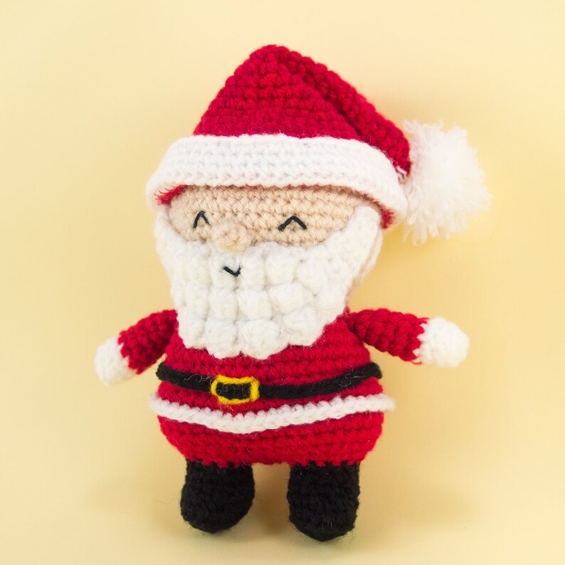 Pode incluir: Um boneco de Papai Noel de croch&ecirc; com um chap&eacute;u vermelho, barba branca e um traje vermelho e branco. O boneco tem um rosto amig&aacute;vel com olhos pretos e um pequeno sorriso.