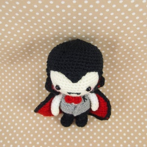 May include: A crocheted vampire doll with a black cape, red lining, and a white face with black eyes and a red bow tie.