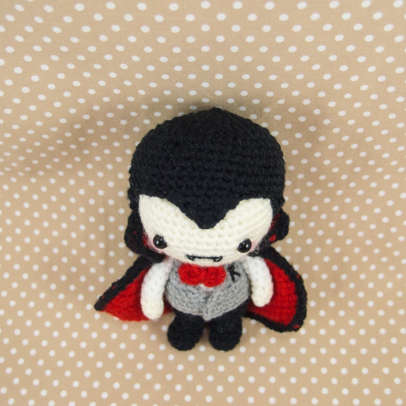 May include: A crocheted vampire doll with a black cape, red lining, and a white face with black eyes and a red bow tie.
