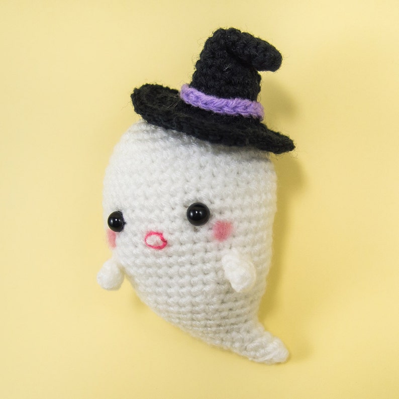 Puede incluir: Un fantasma blanco de crochet que lleva un sombrero de bruja negro con ribete morado. El fantasma tiene ojos negros y mejillas rosadas.