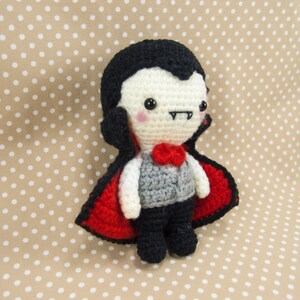 May include: A crocheted vampire doll with a red cape, black hair, and a white face. The doll has a small red bow tie and a gray vest.
