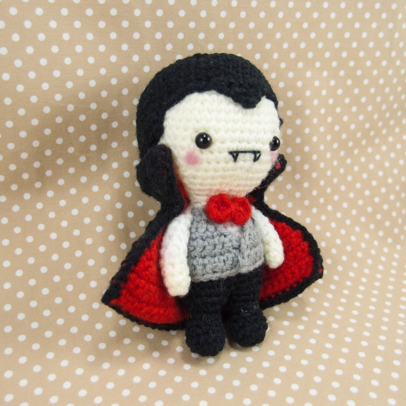 May include: A crocheted vampire doll with a red cape, black hair, and a white face. The doll has a small red bow tie and a gray vest.