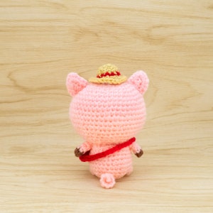 May include: A pink crocheted pig wearing a straw hat with a red band. The pig is holding a red and brown crocheted bag.