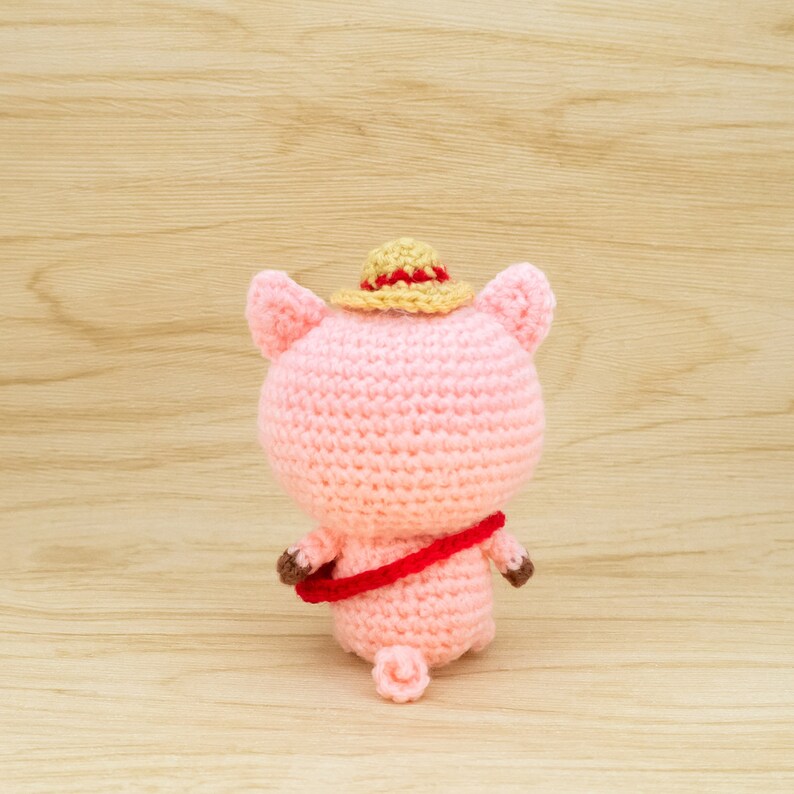 May include: A pink crocheted pig wearing a straw hat with a red band. The pig is holding a red and brown crocheted bag.