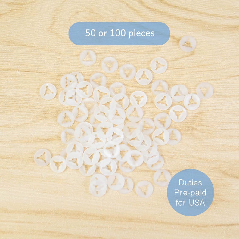 May include: A collection of small, white plastic fasteners with a circular shape and a triangular design in the center. The text "50 or 100 pieces" is visible. The fasteners are scattered on a light wood surface.