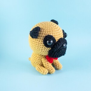 May include: A crocheted pug dog toy with black eyes and a red bow tie. The toy is sitting on a light blue background.
