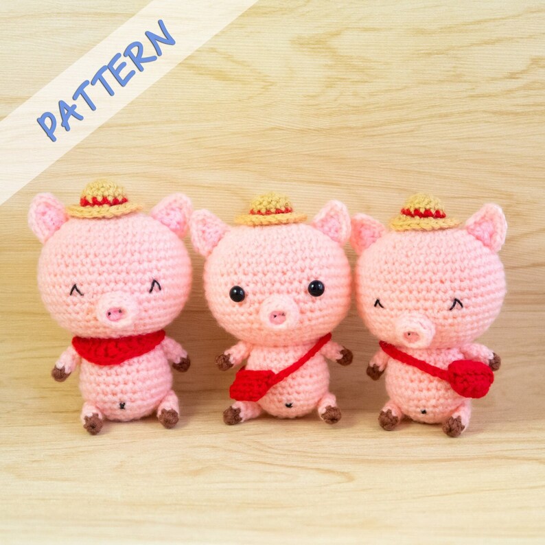 May include: Three crocheted pink pig plushies with brown noses and red accents. Each pig is wearing a straw hat and has a different expression. The pigs are sitting on a wooden surface.