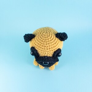 May include: A crocheted pug dog toy with black ears, eyes, and nose. The toy is brown and is sitting on a light blue background.