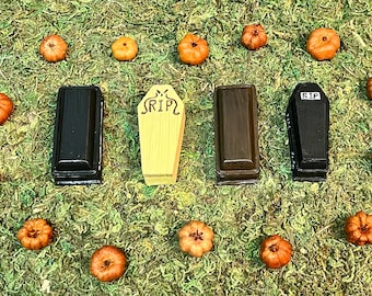 May include: A Halloween-themed arrangement featuring miniature coffins and pumpkins on a bed of green moss. The coffins are black, brown, and yellow, with one displaying "RIP". The pumpkins are orange.