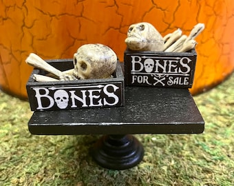 May include: Two black wooden crates with white lettering that says "Bones for Sale" and "Bones" with a skull icon. The crates are filled with miniature white plastic bones and skulls. The crates are sitting on a black wooden table with a pedestal base.