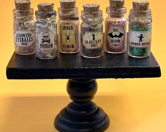 May include: Seven small glass bottles with cork stoppers and Halloween-themed labels on a black tiered cake stand. The labels include "Assorted Eyeballs", "Poison", "Spider Venom", "Graveyard Dirt", "Bat Blood", and "Voodoo Potion".