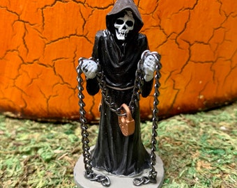 May include: A black skeletal figure wearing a hooded robe and holding two chains with a padlock attached. The figure is standing on a gray base.