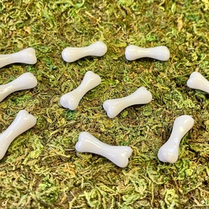 May include: Ten white bone-shaped beads, scattered on a bed of green moss. The beads are smooth and glossy, with a slightly rounded shape.