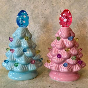 May include: Two ceramic Christmas tree decorations, one light blue and one pink. Each tree has colorful lights and a decorative egg-shaped topper. The trees are glossy and have a retro aesthetic.