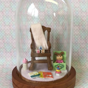 May include: A miniature dollhouse scene under a glass dome. A rocking chair with a white blanket draped over it, a baby bottle, a stack of alphabet blocks, a Cabbage Patch Kids doll, and a stack of children's books are arranged on a pink felt base.
