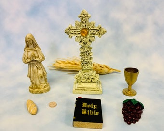 May include: A white ceramic cross with a gold center, a white ceramic statue of the Virgin Mary, a small loaf of bread, a communion wafer, a small book with the words "Holy Bible" on the cover, a small gold chalice, and a bunch of red grapes.