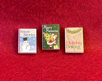 May include: Three miniature books with Christmas themes. The first book is blue and white with the title "Snow Stories" and a snowman illustration. The second book is green and red with the title "Merry Memories" and a Santa Claus illustration. The third book is beige with the title "Holiday Baking" and a holiday baking illustration.