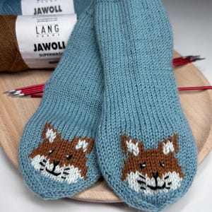 Può includere: Un paio di calzini lavorati a maglia blu chiaro con un disegno di faccia di volpe marrone e bianca. I calzini sono esposti su un vassoio di legno con ferri da maglia e filato sullo sfondo.