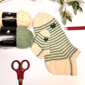 Puede incluir: Un par de calcetines tejidos a mano con rayas verdes y crema, con un diseño de corazón en el tobillo. Dos ovillos de lana y unas tijeras rojas están junto a los calcetines. El hilo está etiquetado como "1 Class Sock Yarn" y "1 Class Cashmere Sock Yarn".