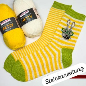 May include: A pair of yellow and white striped socks with green accents, featuring an embroidered bunny wearing glasses. Two skeins of yarn, one yellow and one white, are next to the socks. The socks are handmade.