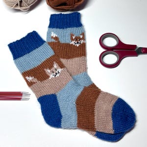 May include: A pair of hand-knitted socks with a fox design. The socks feature blue, light blue, brown, and tan stripes. The fox design is white with black accents. Knitting needles, scissors, and yarn are also visible.