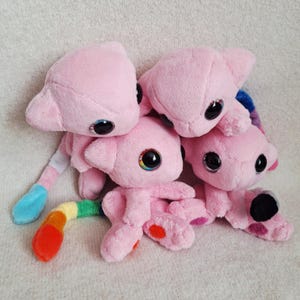 May include: A group of three soft, light pink plush toys with large, dark, reflective eyes. Each creature features pointed ears and a rounded head. One plush has a long, segmented tail with vibrant rainbow-coloured sections including red, orange, yellow, green, and blue.