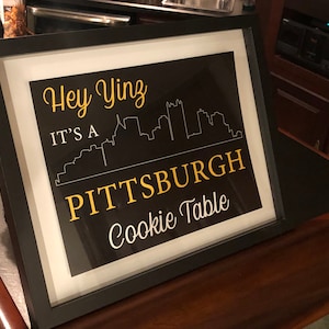 May include: A black framed sign with a white cityscape silhouette and the text "Hey Yinz It's a Pittsburgh Cookie Table" in yellow.