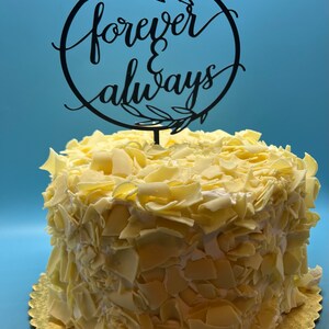 May include: White cake decorated with white chocolate shavings and topped with a black cake topper that reads "forever & always" in a cursive font.