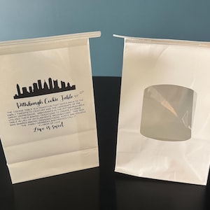 May include: Two white paper bags with a clear plastic window and a skyline graphic. One bag has text that reads "Pittsburgh Cookie Table" and "Love is sweet."