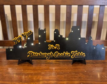 Può includere: Un ritaglio in legno nero dello skyline di Pittsburgh con scritta gialla che recita "Hey Yinz It's A Pittsburgh Cookie Table".