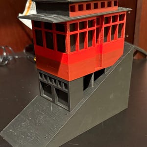 May include: A 3D printed model of a red and black building with a sloped roof and multiple windows. The building is on a black, sloped base.