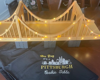 Può includere: Un ponte giocattolo giallo con luci, una borsa nera con il testo "Hey Yinz It's a Pittsburgh Cookie Table" stampato su di essa.