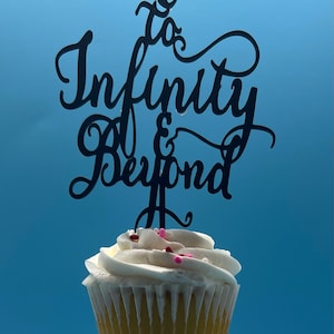 May include: A white cupcake with pink and red sprinkles topped with a black acrylic cake topper that reads "to infinity & beyond".