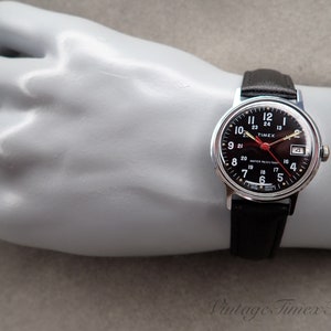 May include: A black-faced Timex wristwatch with a silver case and black leather strap. The watch face has white numerals and a red second hand. The watch is water-resistant.