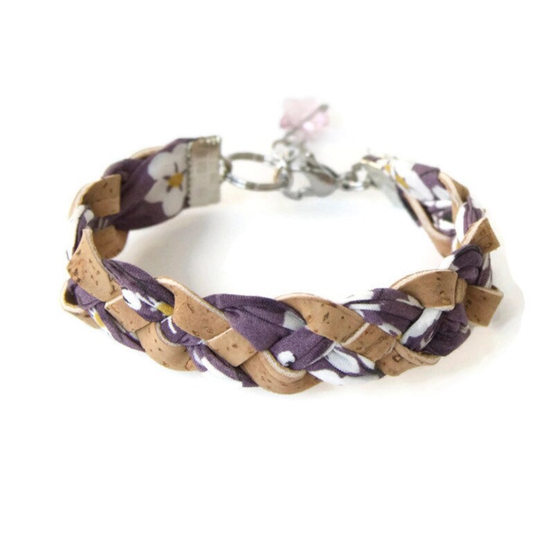 Puede incluir: Un brazalete trenzado hecho de corcho y tela morada con un patr&oacute;n floral blanco. El brazalete tiene un cierre plateado y un dije rosa.