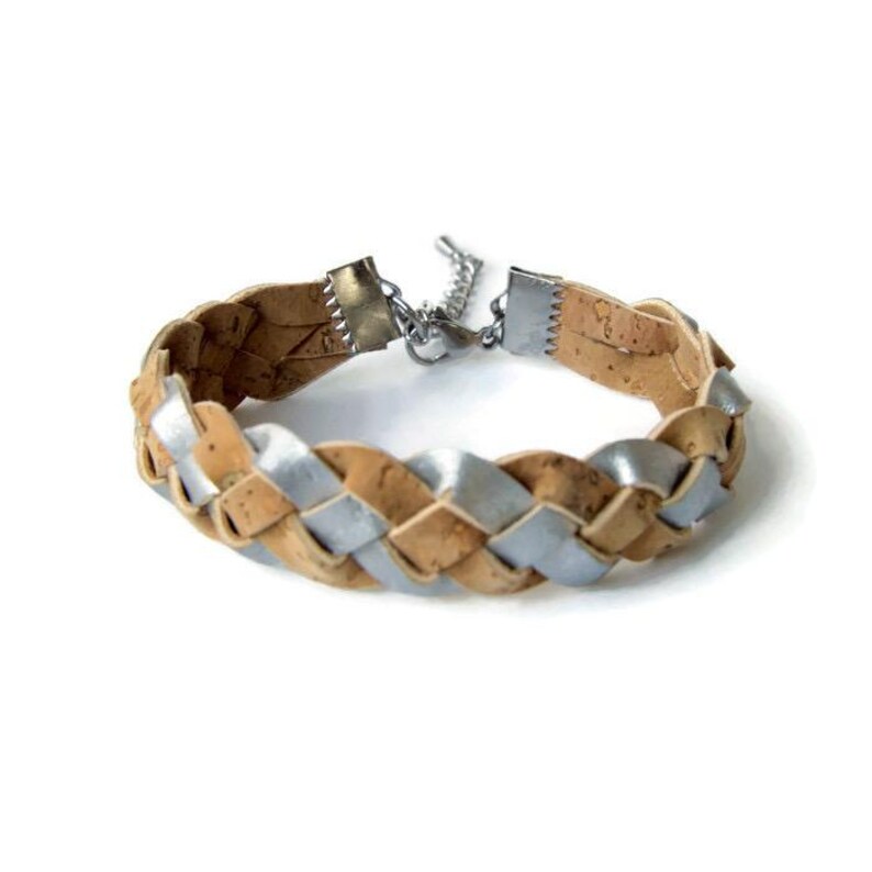 Puede incluir: Un brazalete trenzado hecho de corcho y cuero de color plateado. El brazalete tiene un cierre de color plateado.