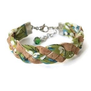Puede incluir: Un brazalete trenzado hecho con tela verde y corcho. La tela tiene un patr&oacute;n floral con flores blancas y detalles azules. El brazalete tiene un cierre plateado y un dije de cristal verde.