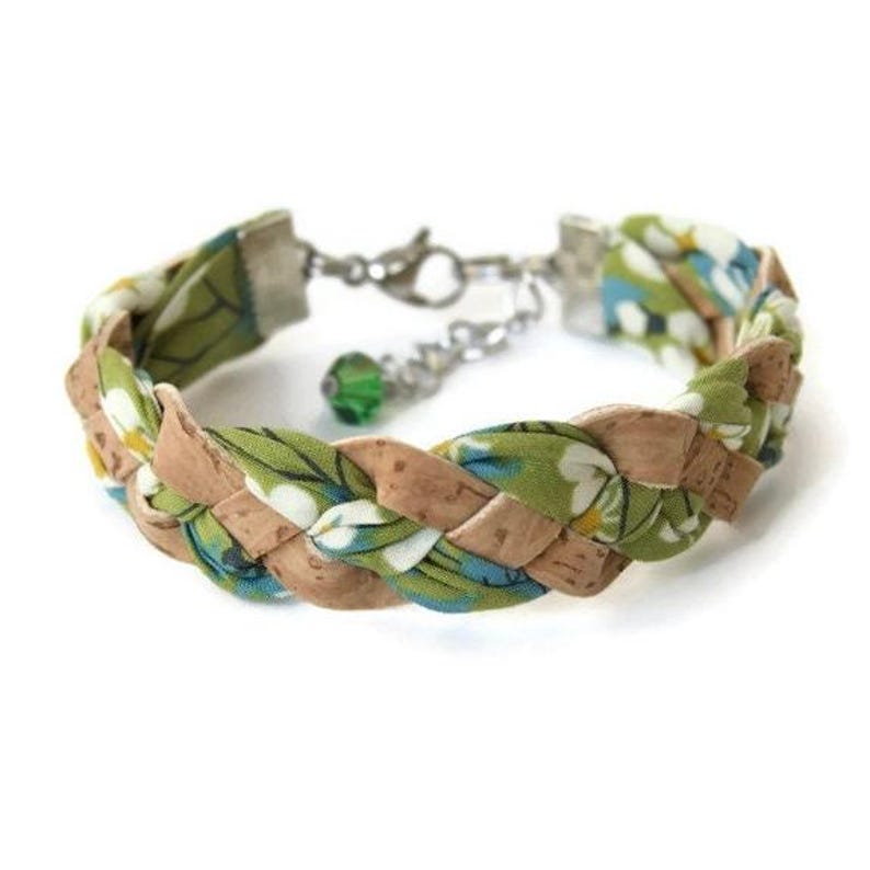 Puede incluir: Un brazalete trenzado hecho con tela verde y corcho. La tela tiene un patr&oacute;n floral con flores blancas y detalles azules. El brazalete tiene un cierre plateado y un dije de cristal verde.