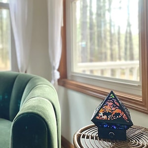 The Wonderland Shadow Lamp staged in a living room as home decor. It is off, in a well lighted room on a wicker table next to a green chair and in front of a window.