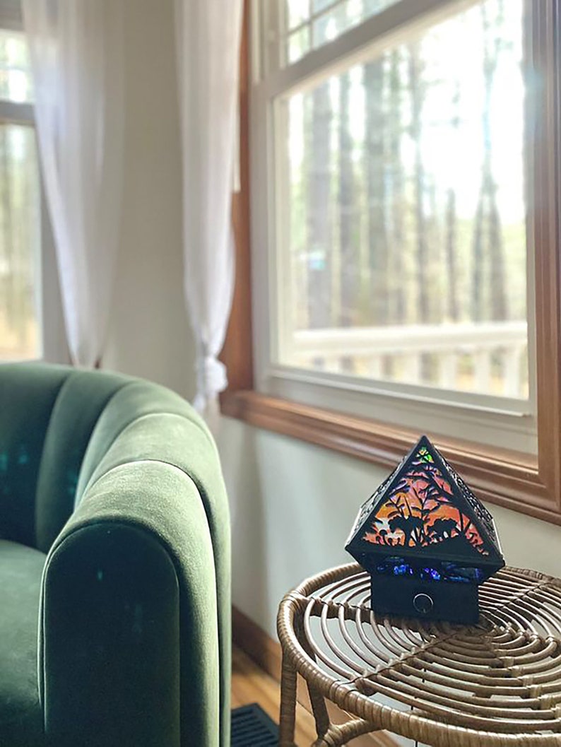 The Wonderland Shadow Lamp staged in a living room as home decor. It is off, in a well lighted room on a wicker table next to a green chair and in front of a window.