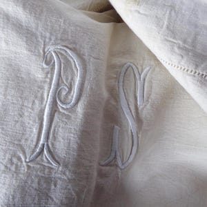 May include: Close-up of off-white linen fabric with embroidered initials "P" and "S" in white thread. The fabric is folded, showing texture and a decorative stitched edge. The embroidery is detailed and elegant.
