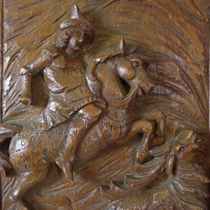 Könnte beinhalten: Eine Holzschnitzerei, die einen Ritter auf einem Pferd zeigt, der einen Drachen tötet. Der Ritter trägt eine Rüstung und einen Helm, und das Pferd bäumt sich auf seinen Hinterbeinen auf. Der Drache liegt am Boden, mit dem Kopf zurückgedreht, um dem Ritter zu begegnen.