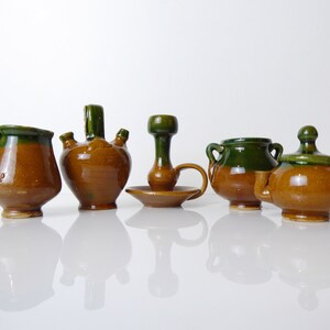May include: A collection of five handcrafted ceramic pieces. The pottery features a brown base with green accents. Items include a small pitcher, a multi-spout vessel, a candlestick holder, a small handled pot, and a lidded jar. The pieces are arranged on a reflective surface.