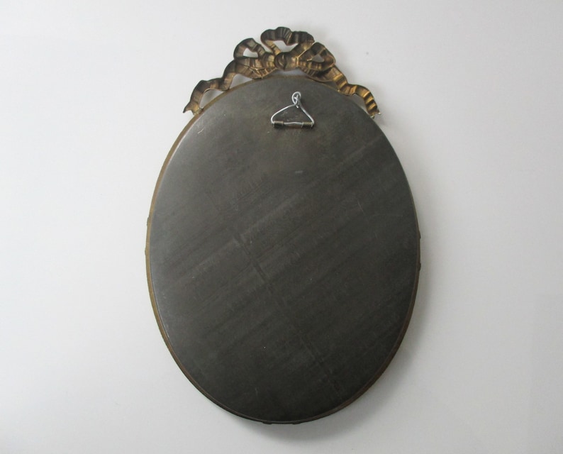 May include: Oval-shaped mirror with a gold-colored ornate frame. The frame features a decorative ribbon design.