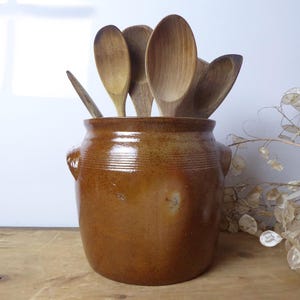 May include: A brown ceramic utensil pot with two small handles, filled with wooden spoons. The pot has a glossy finish and is set on a wooden surface. The spoons are of various sizes.