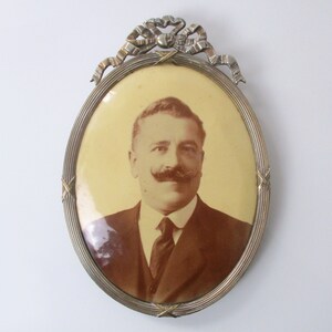 May include: An oval-shaped silver picture frame with a decorative bow at the top. The frame holds a sepia-toned photograph of a man with a mustache wearing a suit and tie.