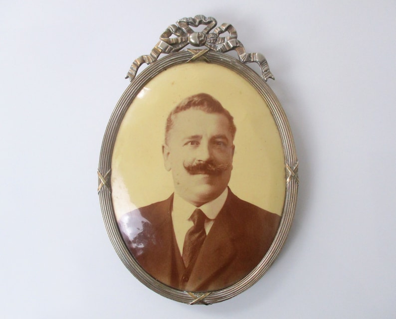 May include: An oval-shaped silver picture frame with a decorative bow at the top. The frame holds a sepia-toned photograph of a man with a mustache wearing a suit and tie.
