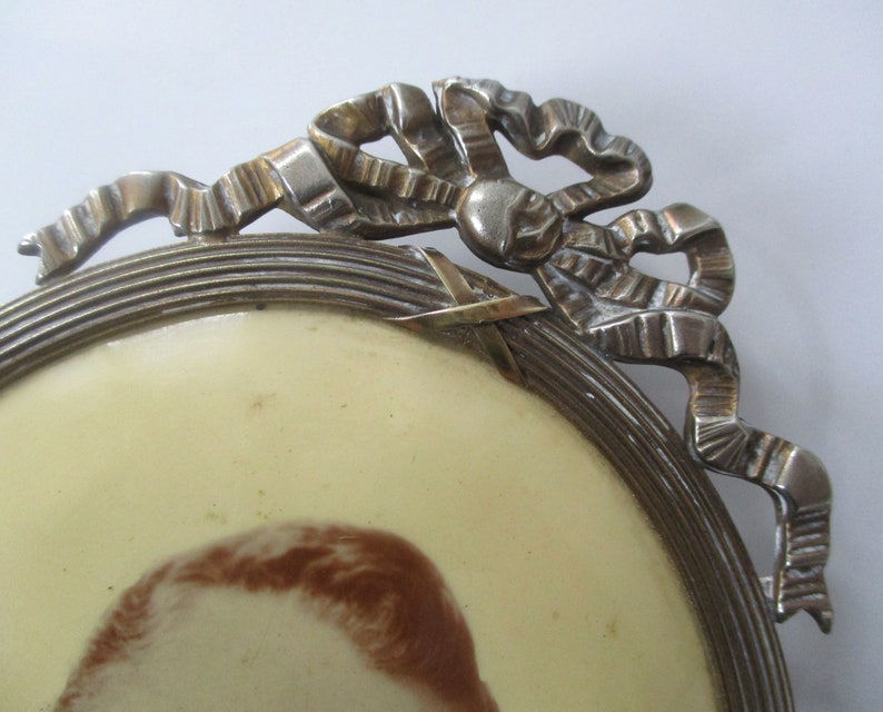 May include: A silver oval picture frame with a decorative bow design. The frame has a raised edge and a smooth, polished finish.